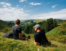 A couple sitting on a grass bank looking at the mountainous views beyond.
