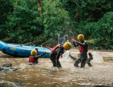 Group of people in safety gear splashing in a river with their rafting boat behind them.