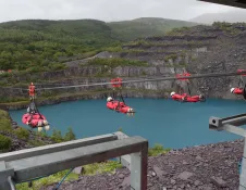 Group of people on a zip wire, flying over a blue reservoir and slate landscape.