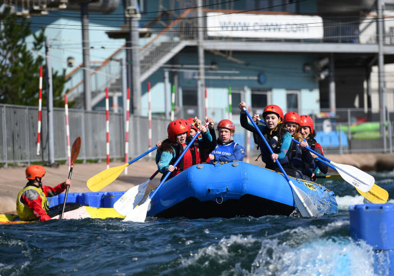 A group kitted out in helmets and lifej ackets using their oars to take on the rapids in a blue dinghy.