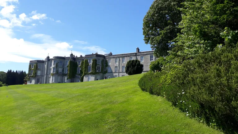 A grand house at the top of a lawned hill framed by trees.