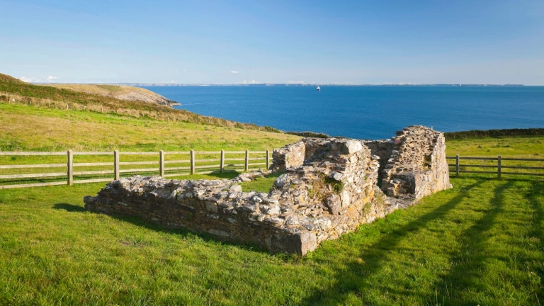 The ruins of St Non's Chapel with a wooden fence surrounding it overlooking the sea.