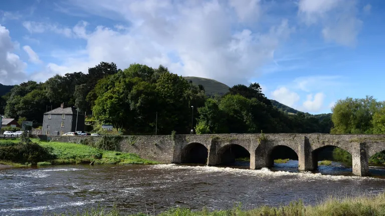 A stone bridge with a river flowing under it. surrounded by trees and a mountain.