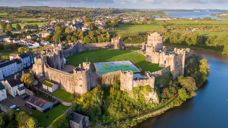 An aerial shot of a large castle with towers alongside the banks of a pond.