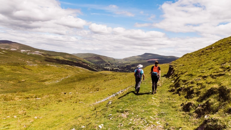 Two walkers on a path heading towards dramatic mountains.