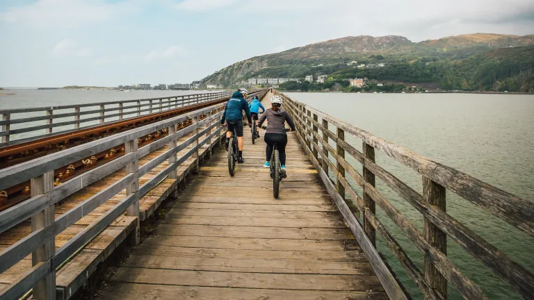 Cyclists on a bridge alongside a railtrack, heading across the estuary toward the coast.