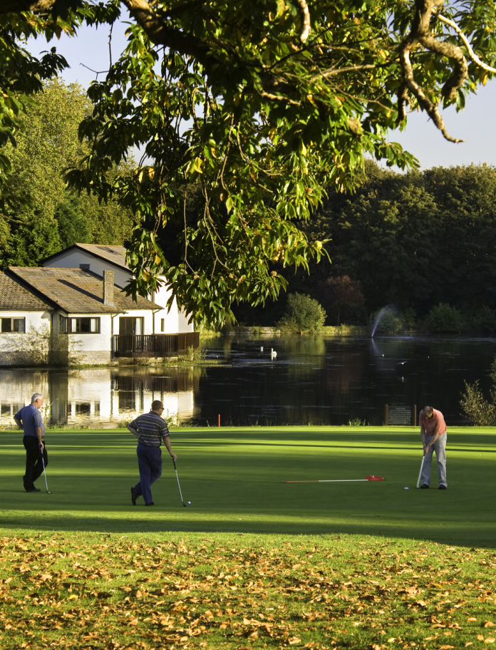 Golfers on the green in front of a white house reflecting on the lake.