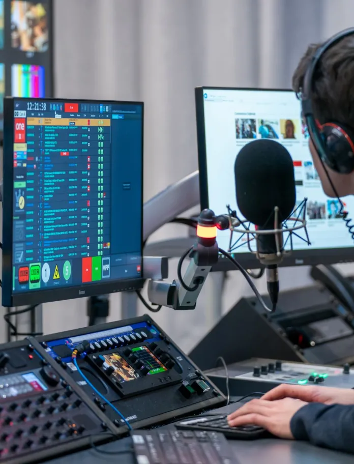 A presenter wearing headphones at a mic and mixing desk in a radio studio.