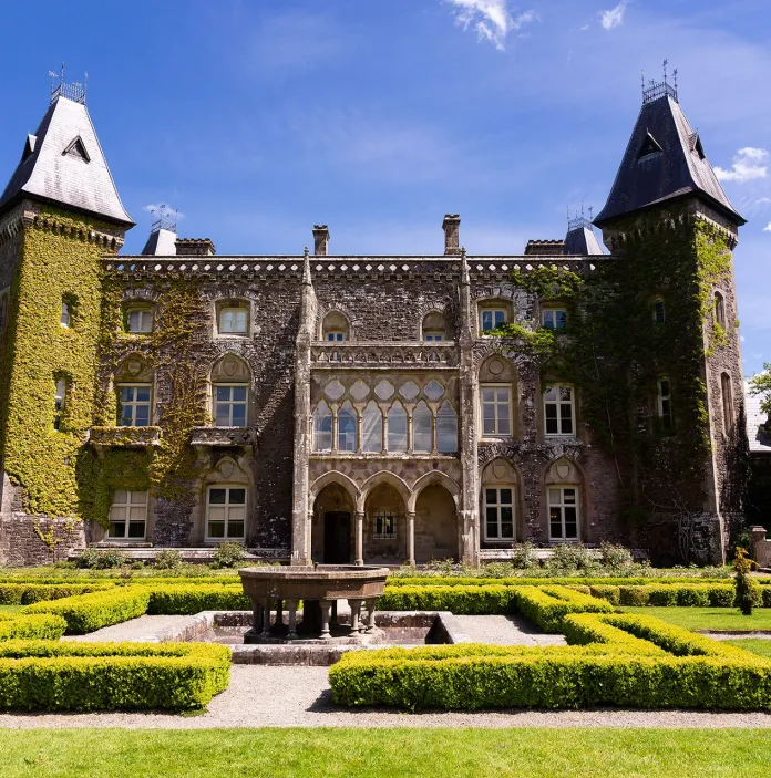 A grand house, covered in ivy with hedged gardens and fountain in the foreground.