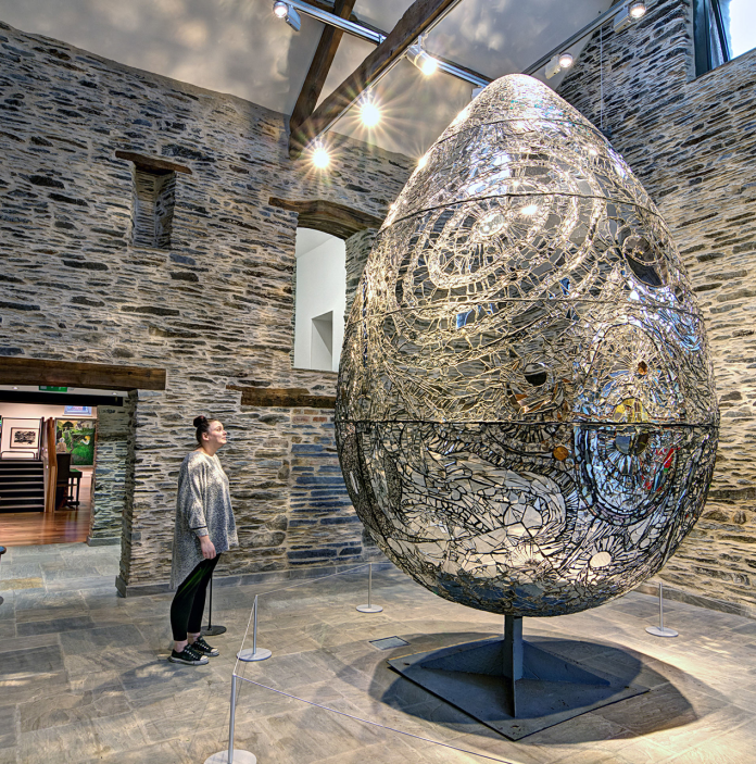 Giant egg sculpture with woman stood looking at it inside a stone wall room.