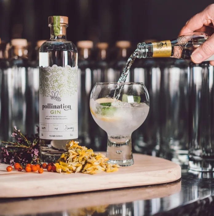A bottle of tonic being poured into a glass of gin on a board showing the gin bottle and ingredients.