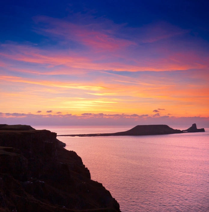 Cliffs in the sea which look like a dragon with a colourful sunset beyond.