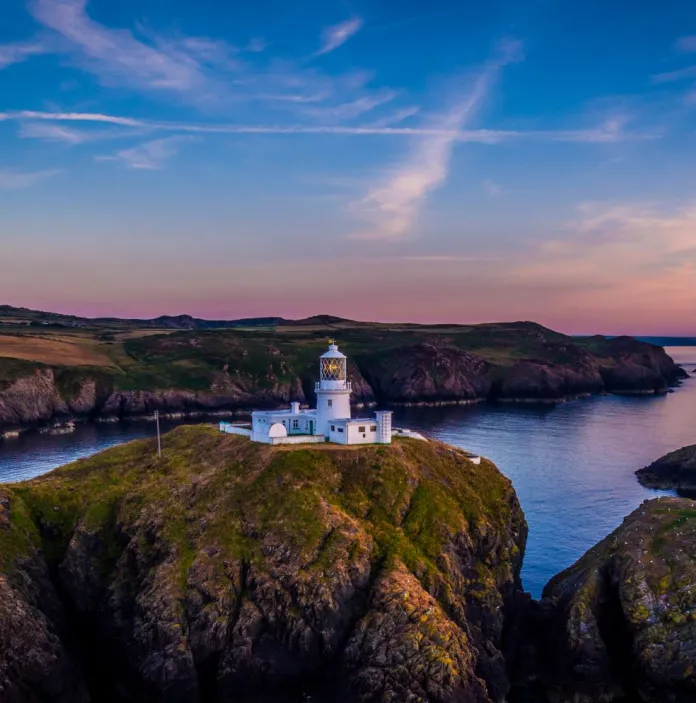 Lighthouse on rocks surrounded by a dramatic coast and sunset.