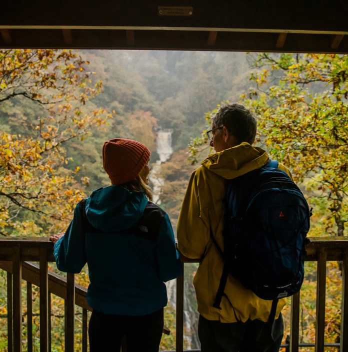 A couple on a viewing platform looking at a waterfall surrounded by autumnal trees.