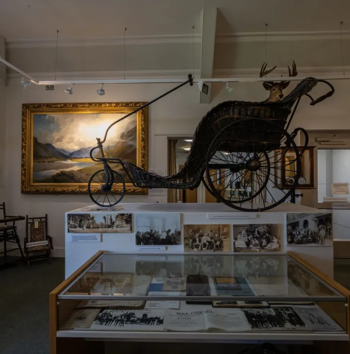An old wicker wheelchair on display with other artefacts in a museum.