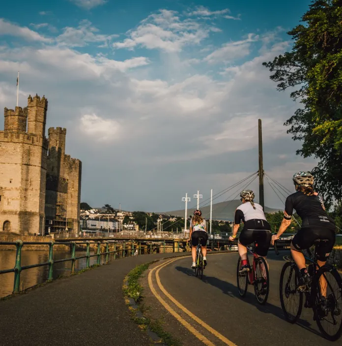 Cyclists wearing safety gear on a road heading alongside a river toward a castle.