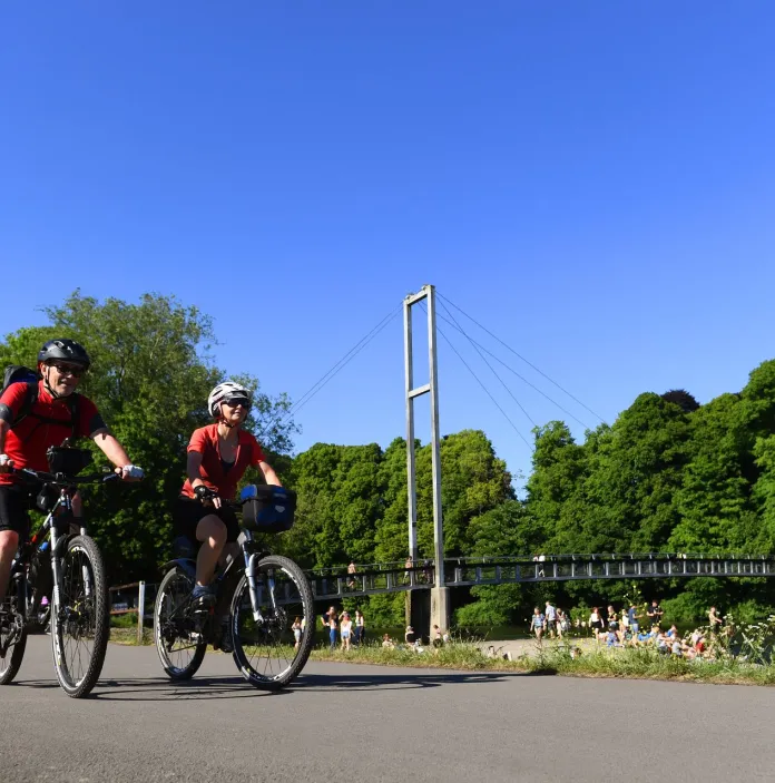 Cyclists wearing safety gear in a park with a footbridge over the water in the background.