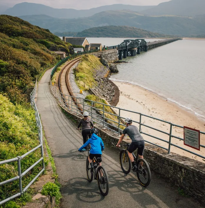 Group of cyclists on a path next to a railway line and beach heading toward a bridge leading over the estuary.