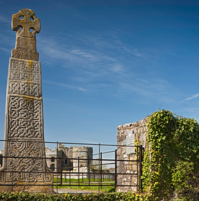 A tall stone celtic cross with carvings with a castle in the background.