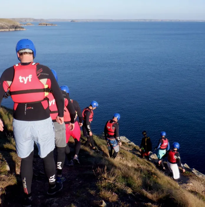 Coasteers wearing protective gear walking down a cliff toward the ocean.