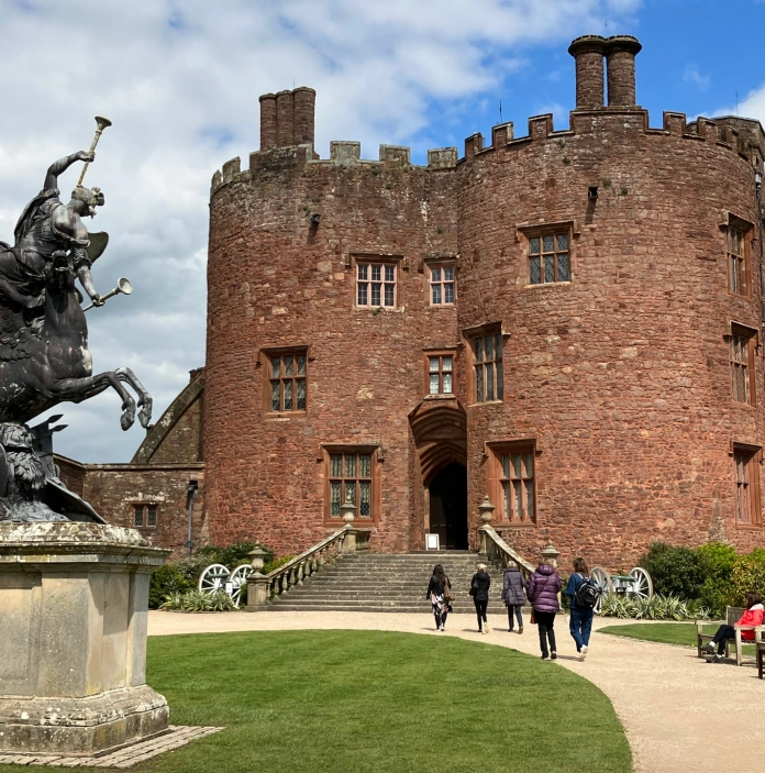 People walking past a statue of a horse and rider toward a grand castle.
