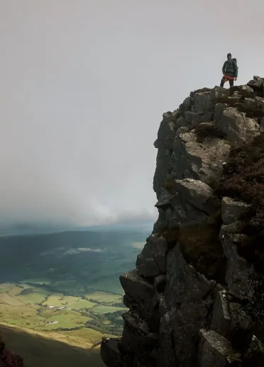 A man standing on a rocky mountain, with views of green pasture beyond.