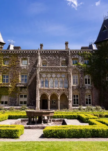 A grand house, covered in ivy with hedged gardens and fountain in the foreground.