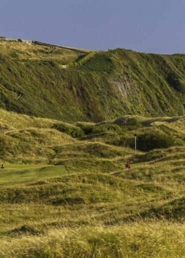 Golfers near 15th green at Royal St David's Golf Club with heath covered cliffs in background.