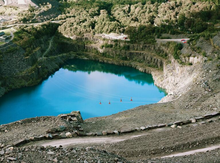 People on ziplines flying over a blue lagoon in a slate quarry.