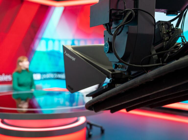 A television camera pointing to a presenter on a sofa.