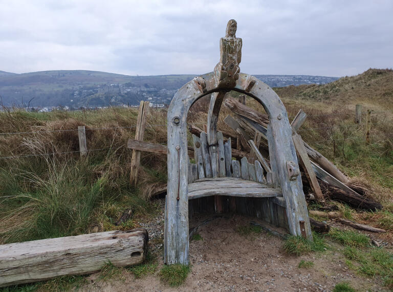 A wooden medieval chair on a walking trail with views of mountains beyond.