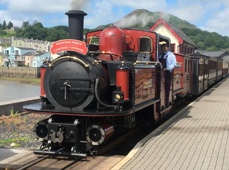 A red steam train setting off from a station next to a harbour.