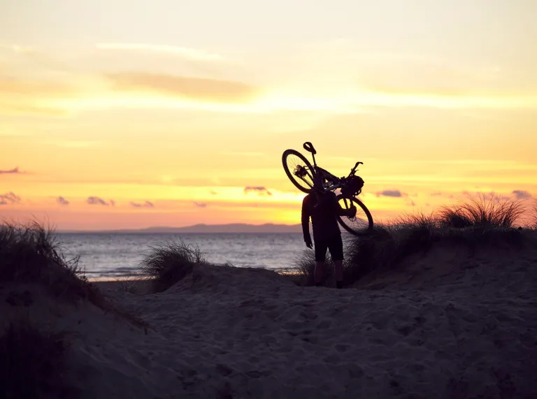 A silhouette of a biker carrying a cycle with a dramatic orange sunset on the coastline.