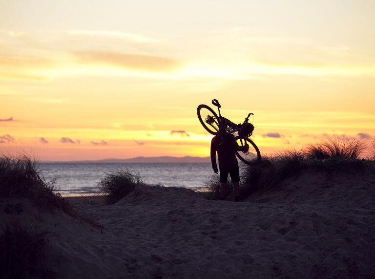 A silhouette of a biker carrying a cycle with a dramatic orange sunset on the coastline.