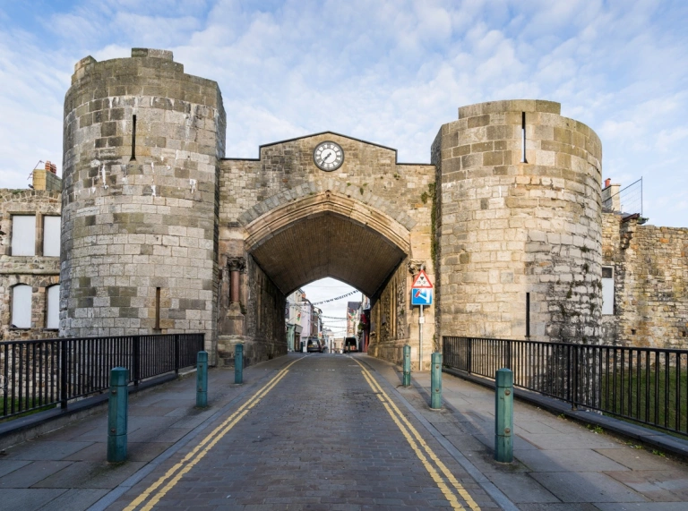 A cobbled road going through an arch in a castle wall leading to a town with turrets either side..