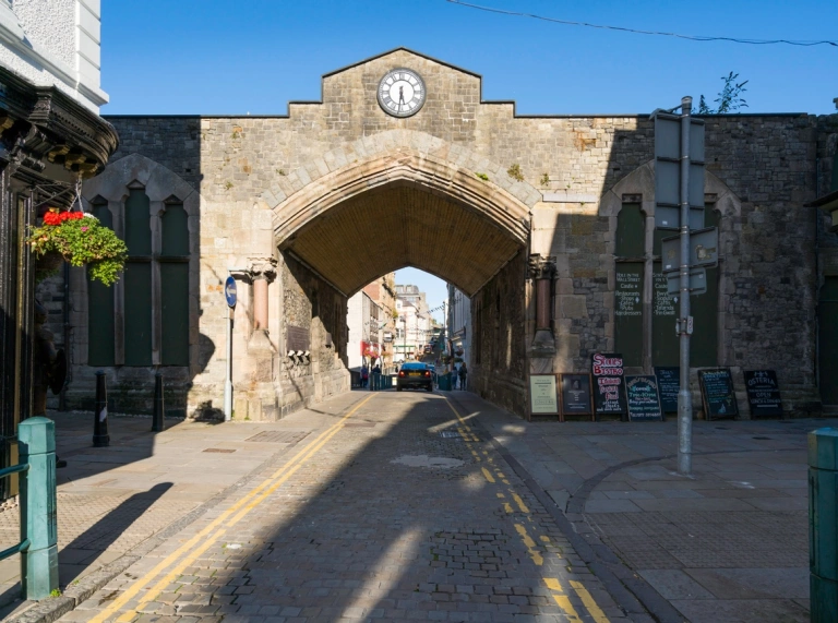 A cobbled road going through an arch in a castle wall leading to a town.