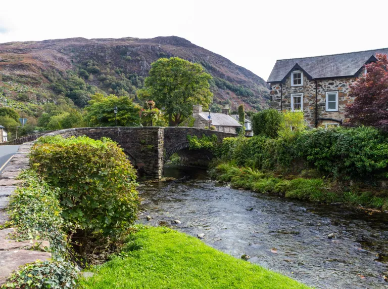 A stone bridge going over a river into a village with mountains in the background.