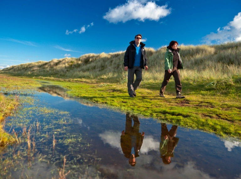 Couple walking by sand dunes with their reflection in a pool of water.