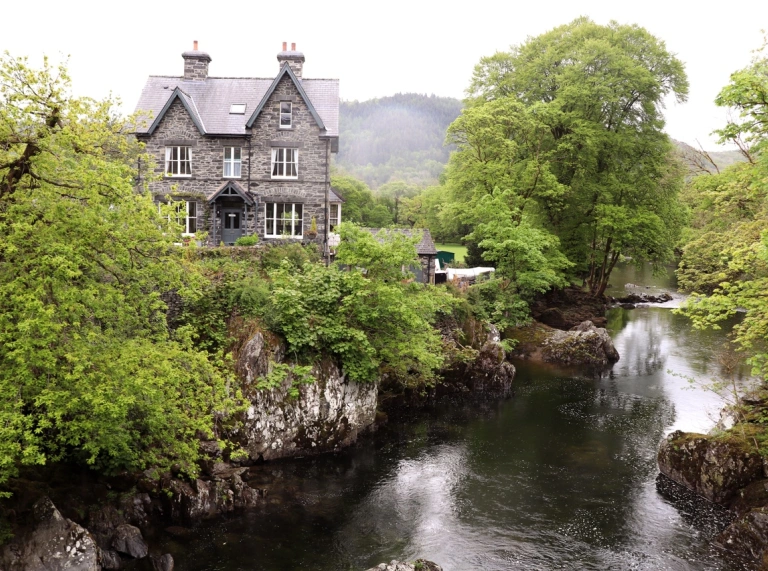 A river running through rock formations and lush green trees with a stone house on the embankment.