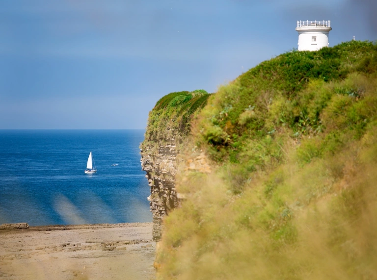 A lighthouse on a cliff with a yacht sailing on the sea below.