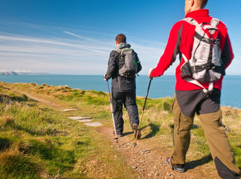 Two walkers wearing appropriate gear hiking along a coast path on a sunny day.