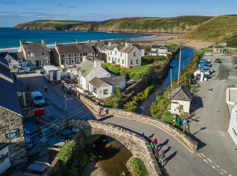 People walking over bridge leading to the village of Aberdaron with the sea in the background.