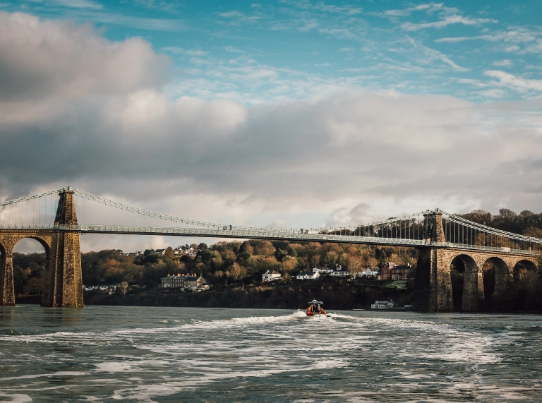Image of the suspension bridge with houses and trees in the distance, and a rib boat on the Menai Strait