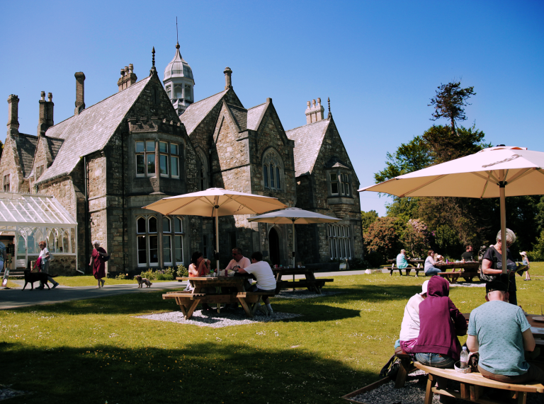 people sat at patio tables with parasols with a manor type building in the background