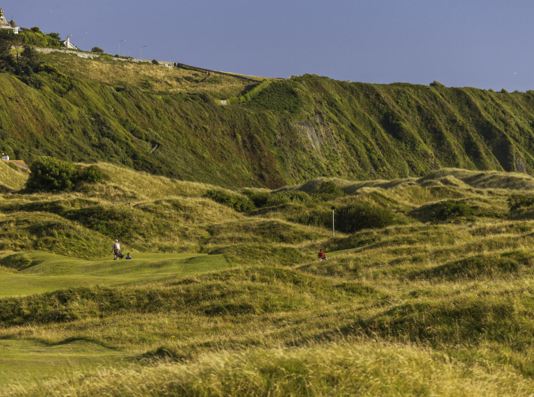 Golfers near 15th green at Royal St David's Golf Club with heath covered cliffs in background.