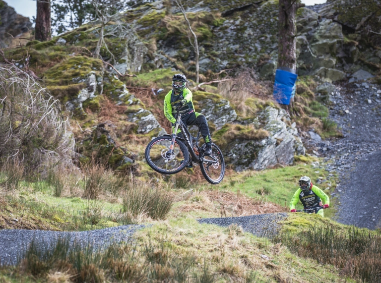 Two bikers on a mountain bike trail, one of which is soaring in the air after a jump.