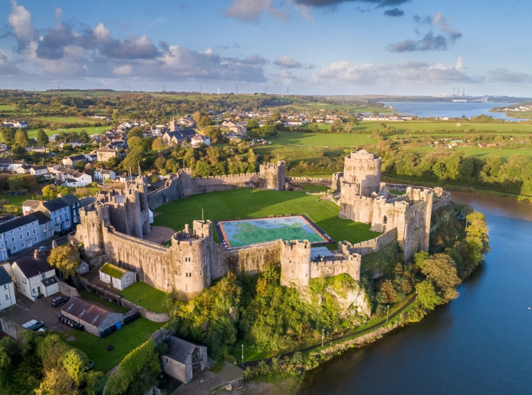 An aerial shot of a large castle with towers alongside the banks of a pond.