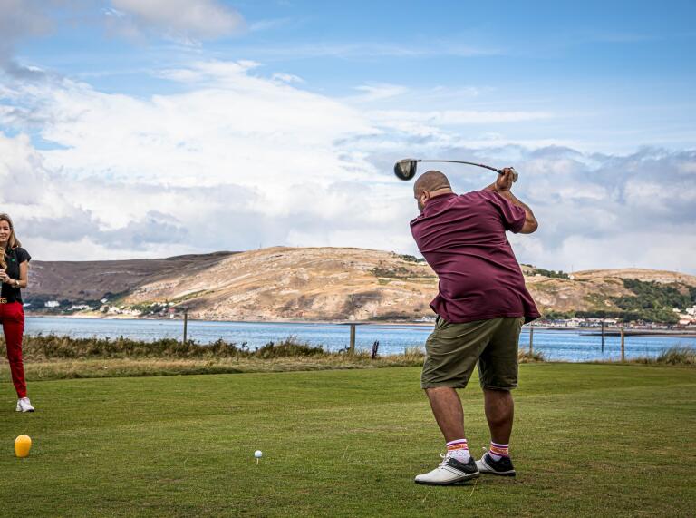 A golfer taking a swing at a ball on a course with views of the sea behind.