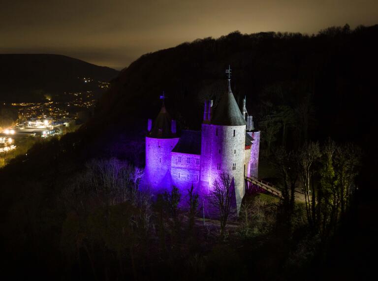A castle colourfully lit up on a hill at night, with views of the village below.