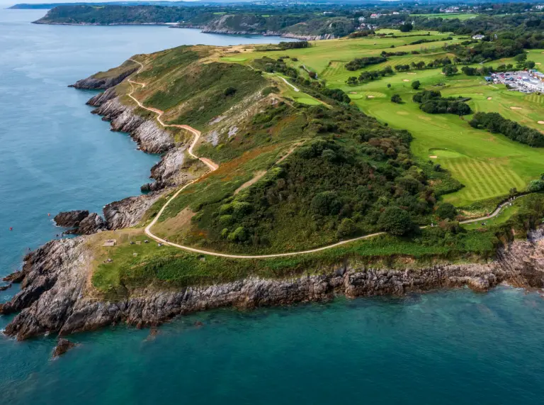 Aerial shot of a coast path on the headland of a bay, with a golf course in the background.